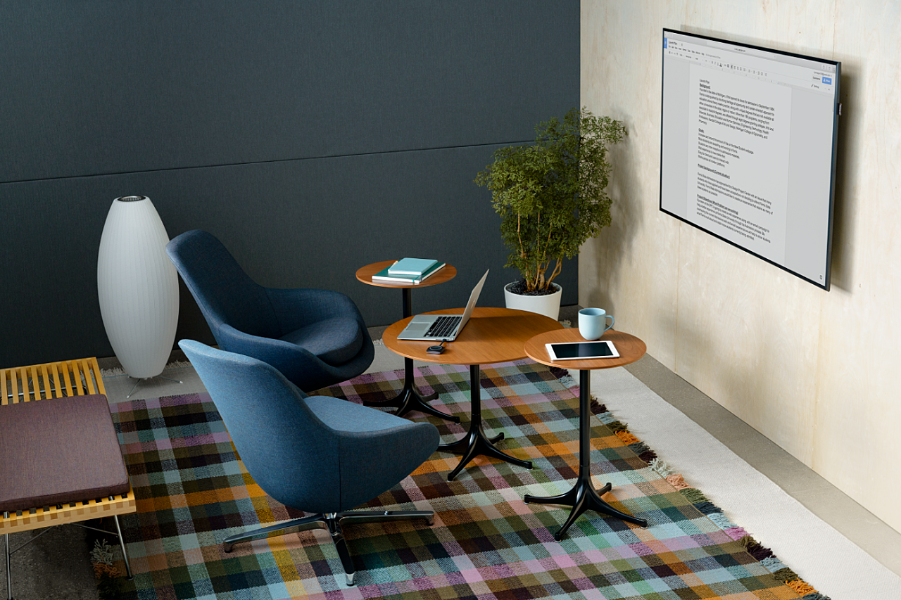 A cozy workspace setup featuring modern blue lounge chairs, wooden tables, a plaid rug, a floor lamp, a potted plant, and a wall-mounted display screen.