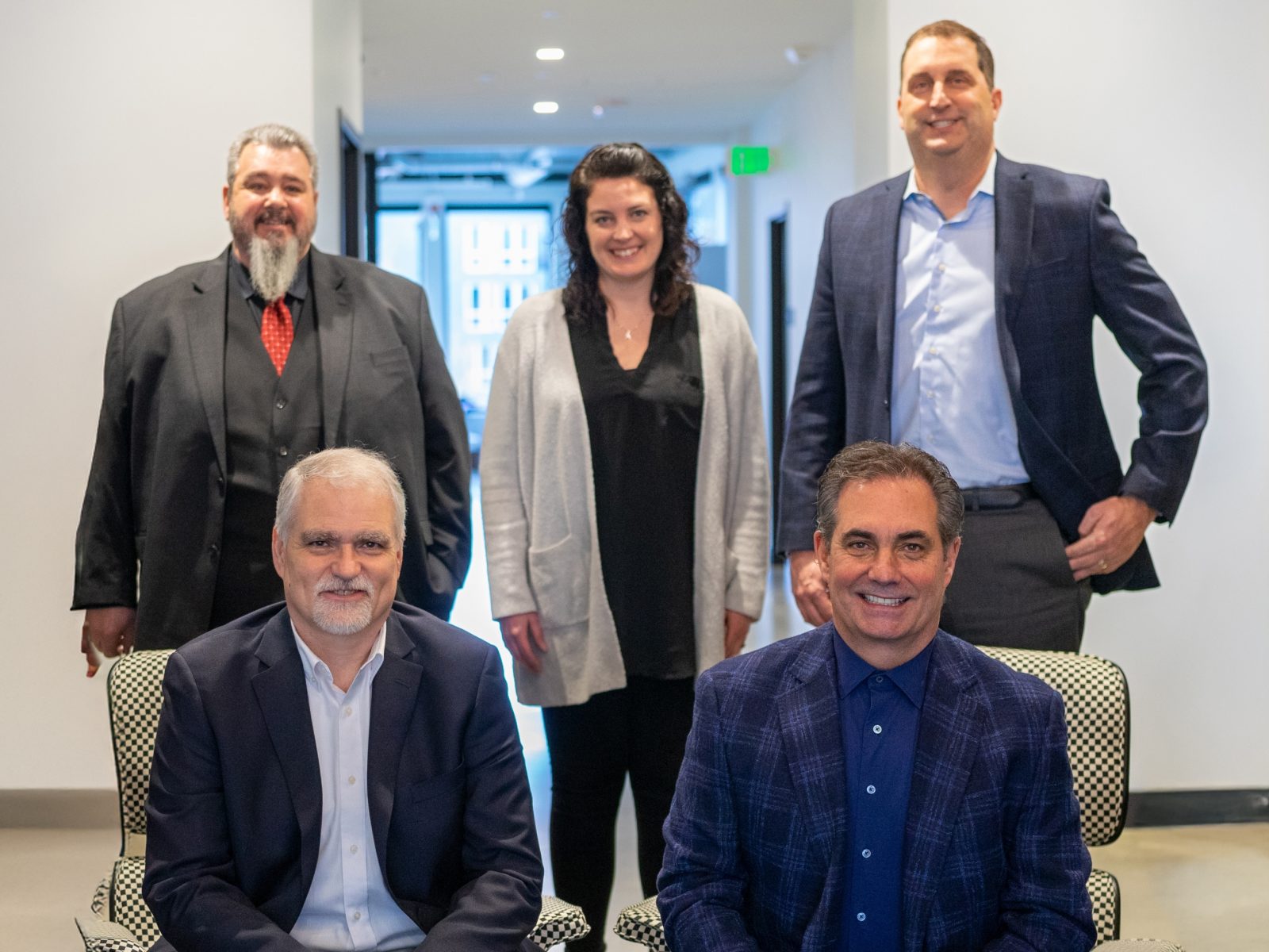 A professional team portrait featuring five individuals in business attire, smiling in a well-lit office corridor, conveying a collaborative atmosphere.