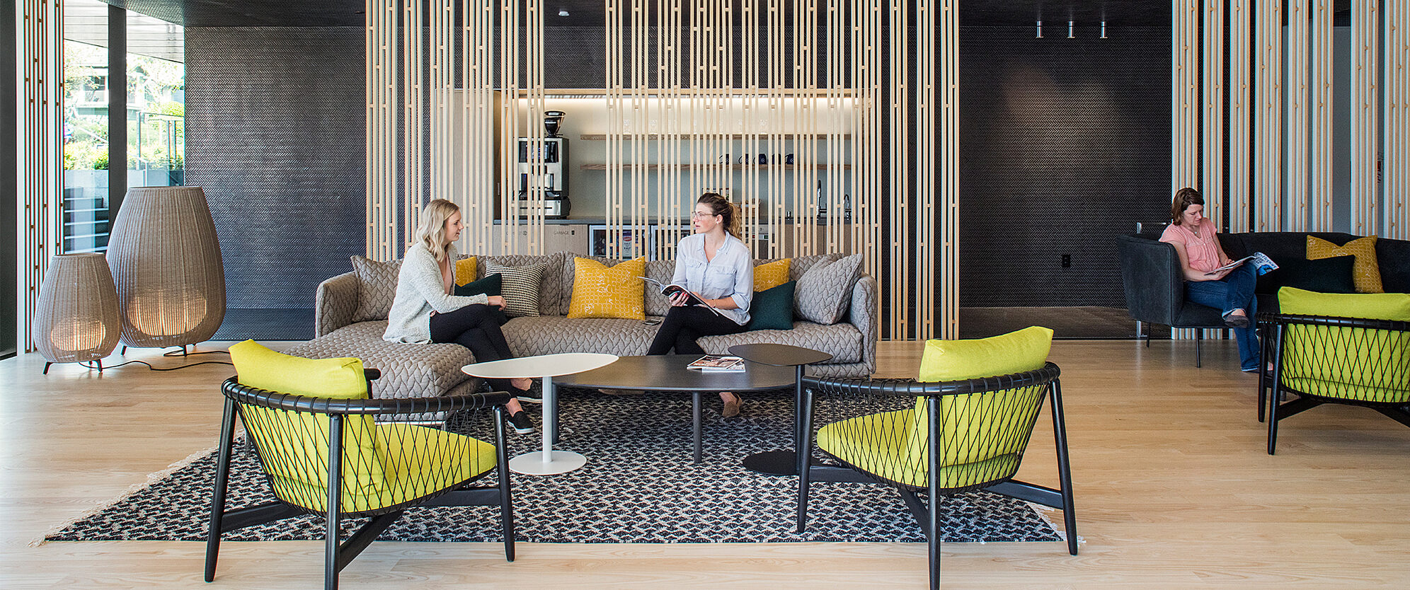 Modern lounge with gray sofas, yellow-accented chairs, and a patterned rug. Two women chat at a round table, while another person relaxes in the background
