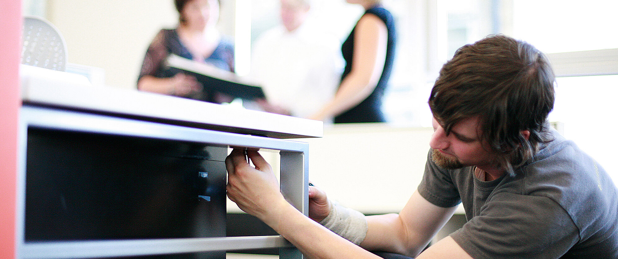 Technician repairing office furniture in a professional workspace with colleagues in the background, highlighting maintenance and support services.