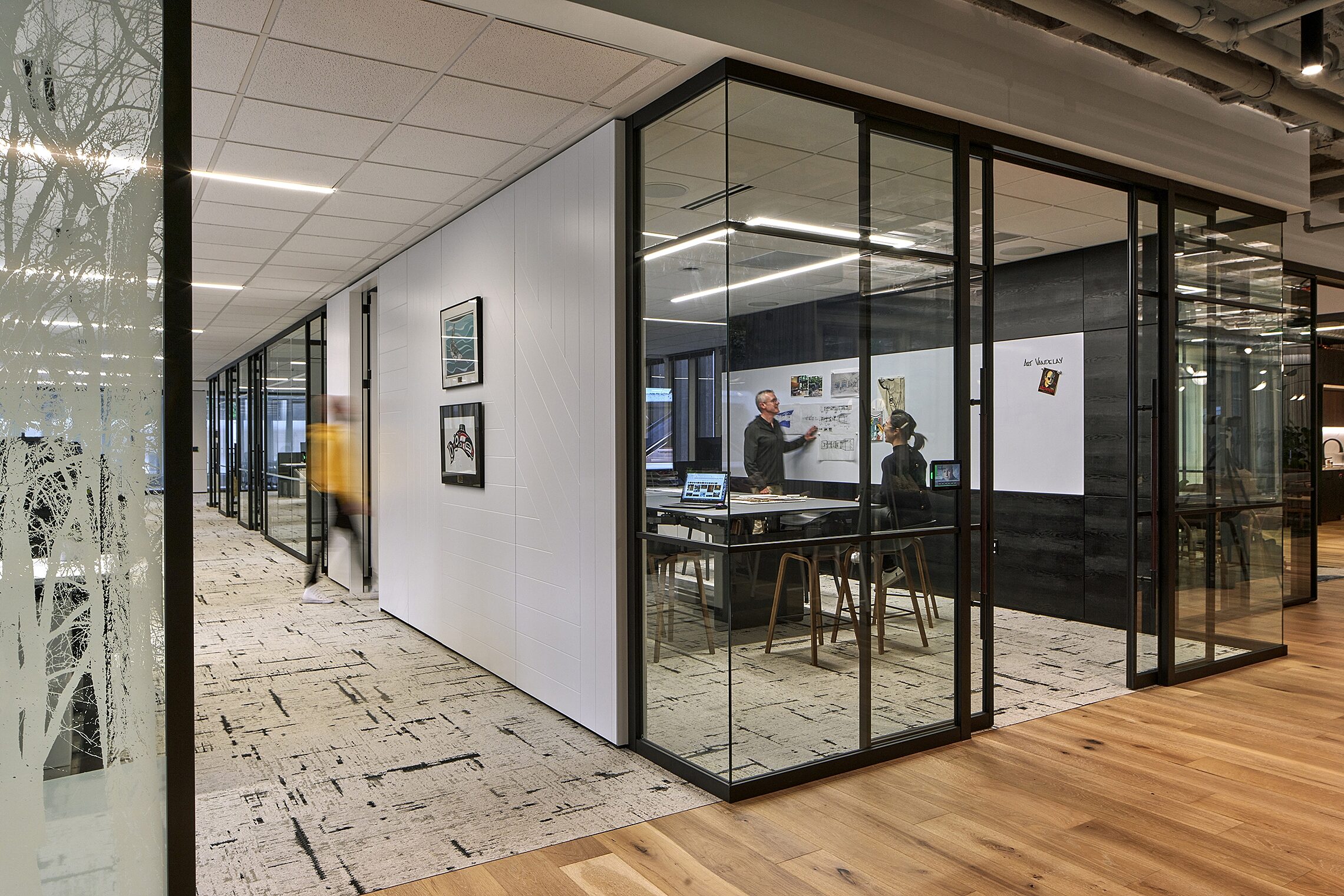 Modern office with glass-walled collaboration rooms. Inside, two people discuss designs on a whiteboard, while a hallway features art and textured floors.