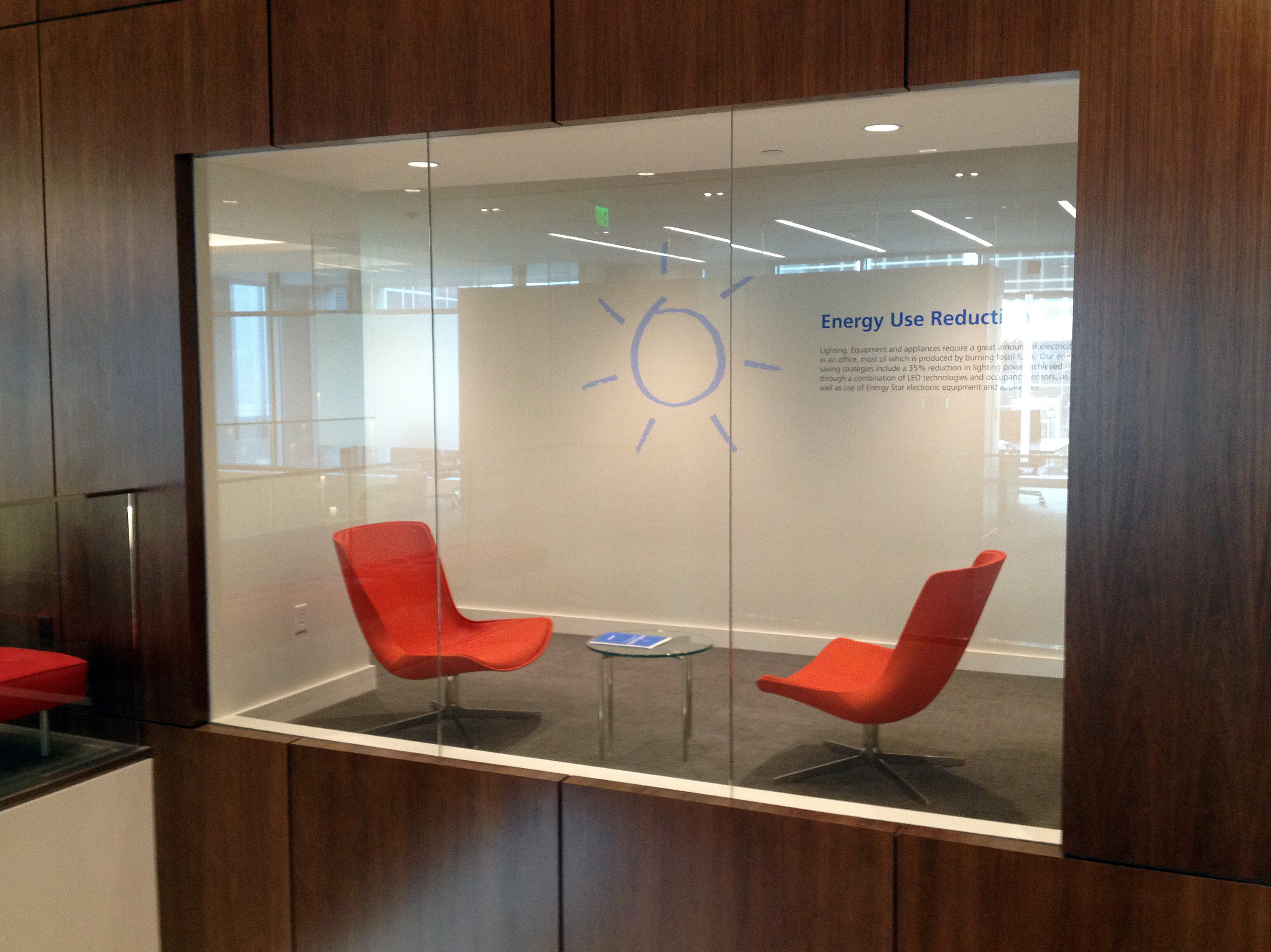 Symbio glass and wood walls enclose a cozy seating nook featuring bright orange chairs, a glass table, and energy use reduction messaging.