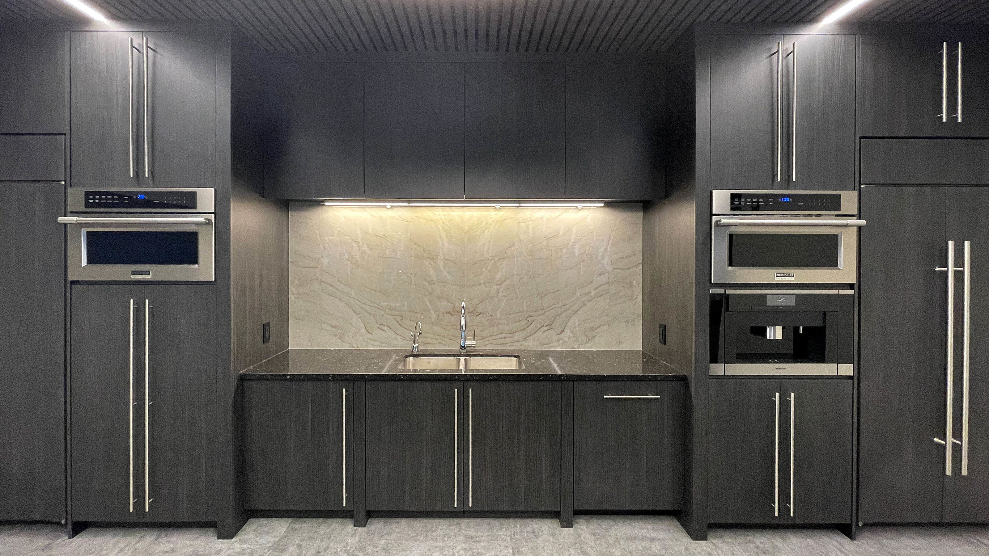 Falkbuilt-designed kitchen with sleek dark cabinetry, built-in appliances, a stainless steel sink, under-cabinet lighting, and a textured backsplash.