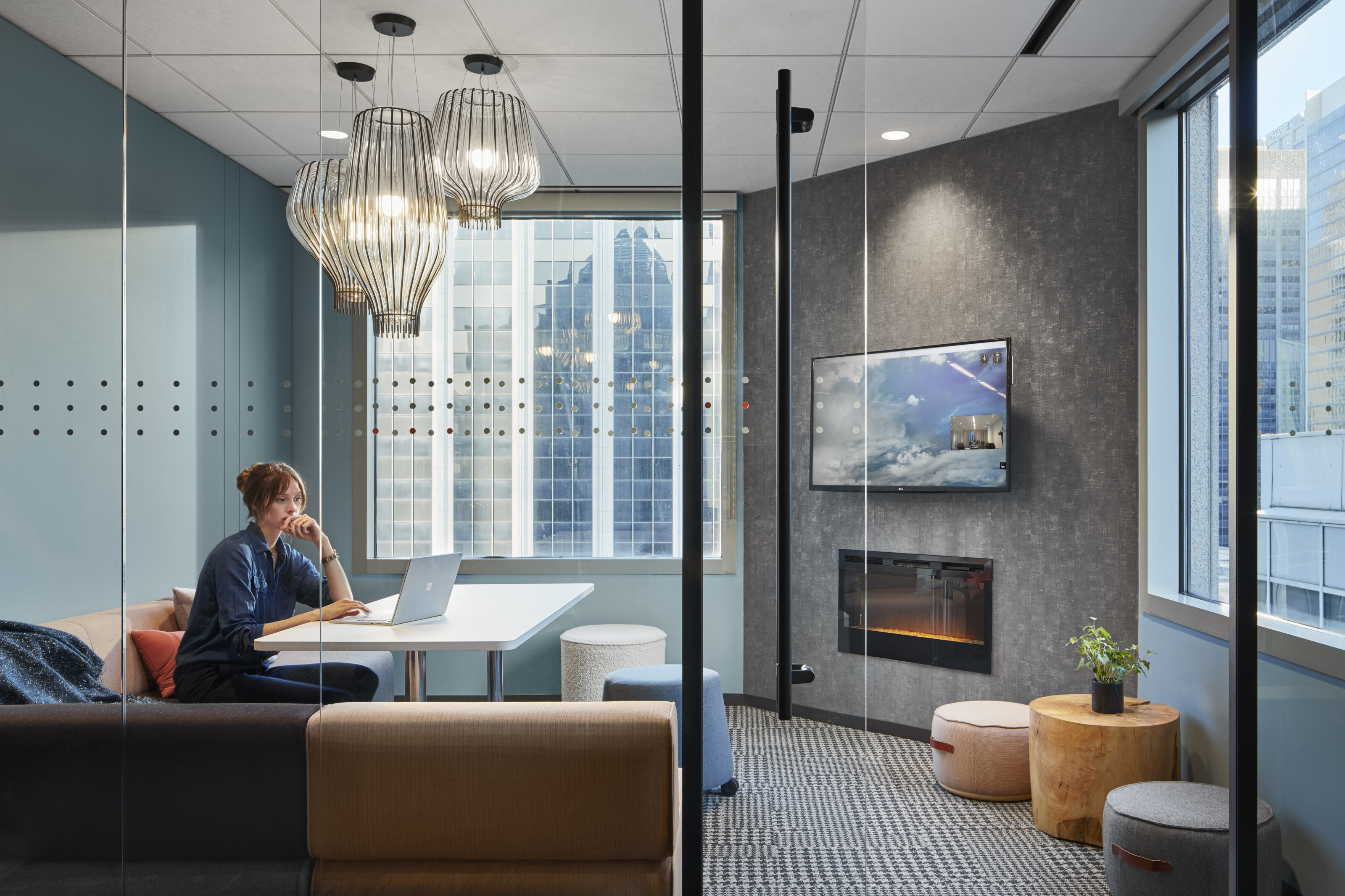 Cozy meeting room with glass walls, a modern fireplace, wall-mounted screen, and pendant lights. A person works on a laptop at a table with city views.