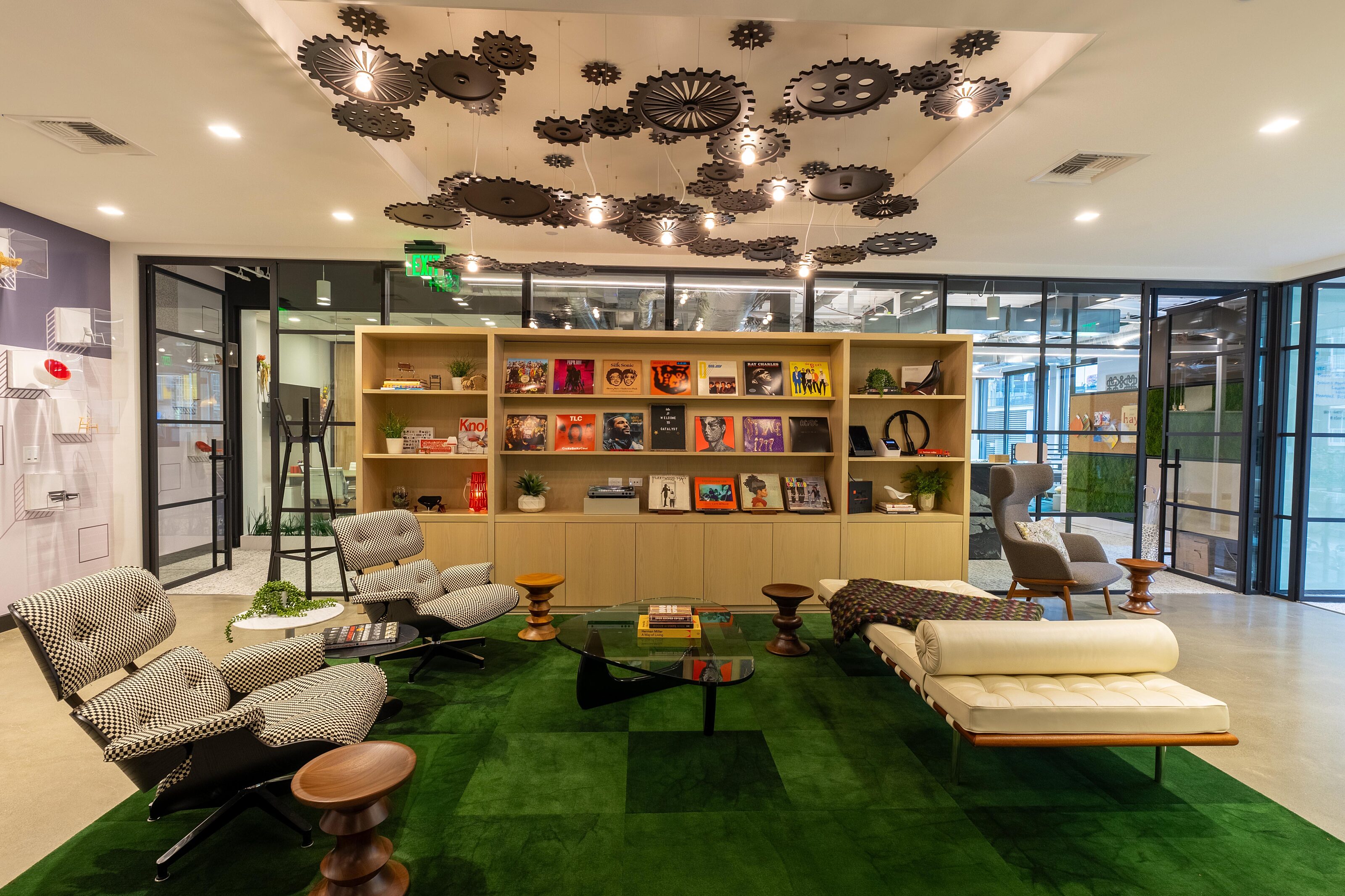 A modern lounge area featuring mid-century furniture, vibrant green carpeting, and a wall display of books and records. Ceiling decor includes gears.