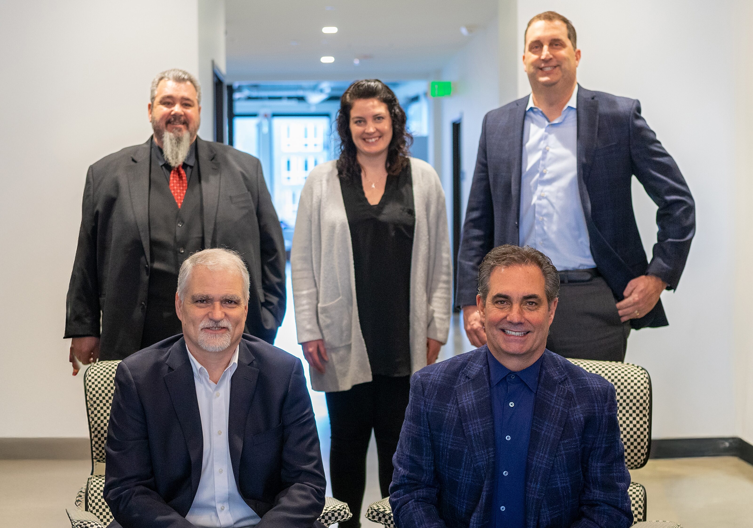 A professional team portrait featuring five individuals in business attire, smiling in a well-lit office corridor, conveying a collaborative atmosphere.