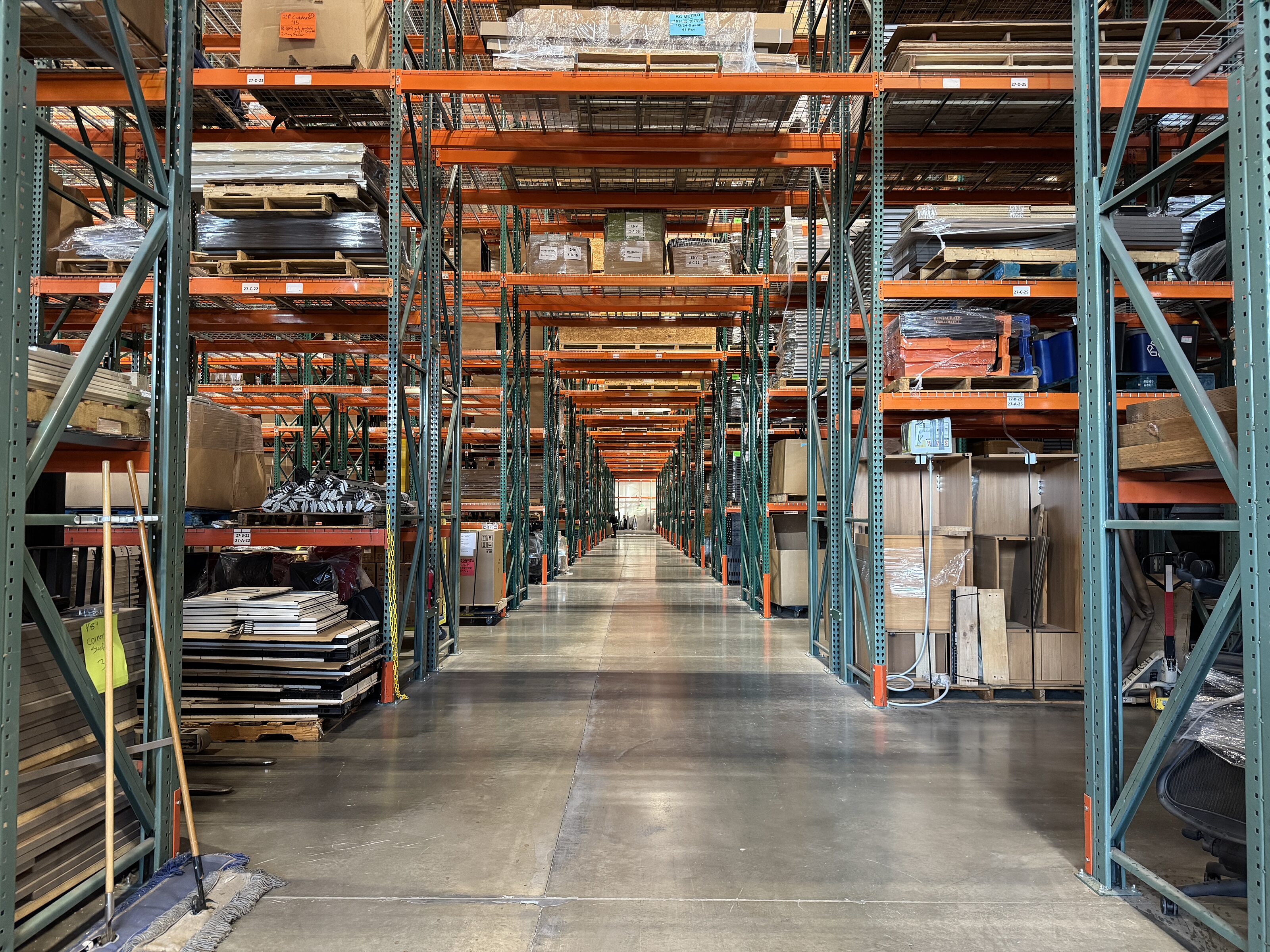A spacious warehouse aisle with tall orange and green metal shelving stocked with various materials and products, neatly organized and stored.