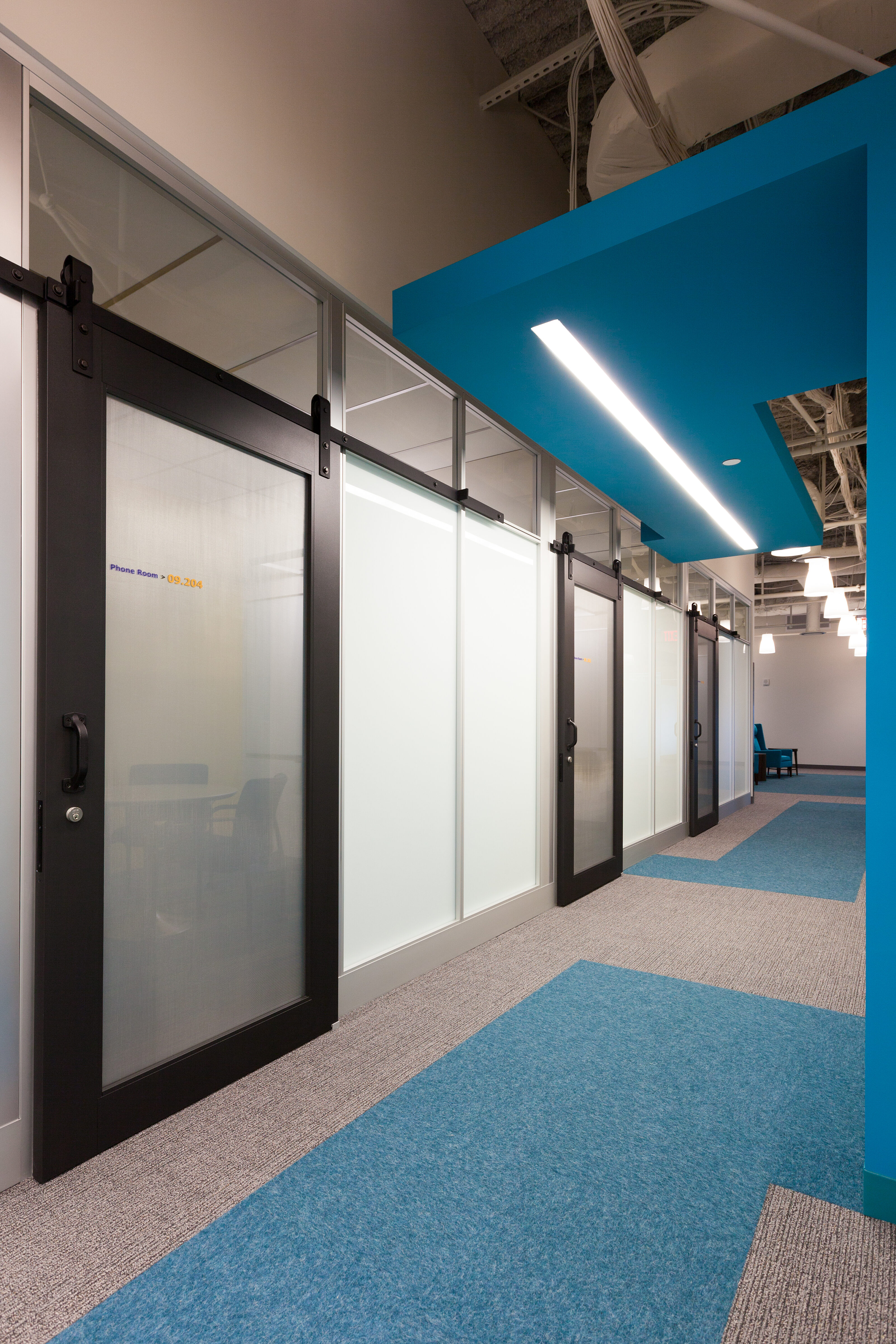 Symbio walls with frosted glass and sleek sliding doors provide modern, private spaces in this office corridor, complemented by vibrant blue accents.
