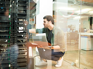 A technician working in a modern server room, holding a laptop and checking hardware connections, reflecting IT infrastructure management in a workspace.