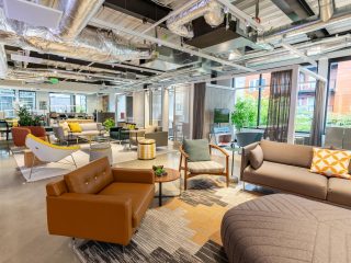 Modern office lounge with colorful seating, sofas, and armchairs arranged on rugs. Large windows and exposed ceilings create an open, inviting workspace.