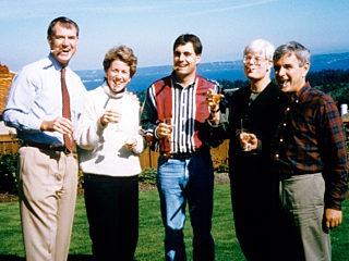 A group of five people standing outdoors on a grassy area, smiling and holding drinks, with a scenic landscape visible in the background.