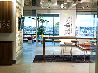 Entryway to Catalyst's Loft space, a large wood communal table and white stools center the space. Floor to ceiling windows show Downtown Seattle skyline.