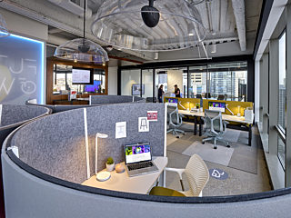 Felted private office pod with seating and workstations in the background in an open office setting. Yellow pops of color accent the workstations.
