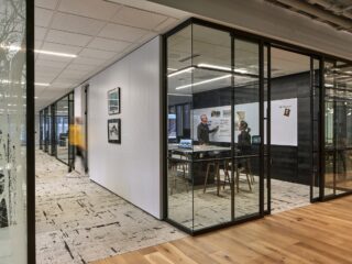 Modern office with glass-walled collaboration rooms. Inside, two people discuss designs on a whiteboard, while a hallway features art and textured floors.