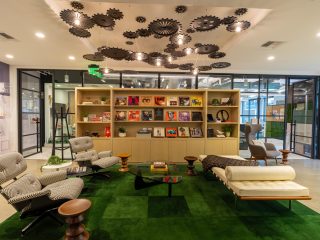 A modern lounge area featuring mid-century furniture, vibrant green carpeting, and a wall display of books and records. Ceiling decor includes gears.