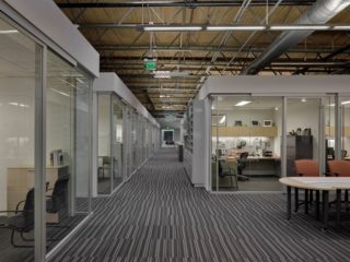 Glass Symbio walls frame private offices and open workspaces along a striped carpeted hallway, with exposed ceilings adding an industrial design touch.