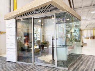 Glass-walled modular office pod with a wood-accented roof and modern interior decor, including seating and decor elements, set in a bright industrial space