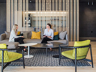 Modern lounge with gray sofas, yellow-accented chairs, and a patterned rug. Two women chat at a round table, while another person relaxes in the background