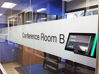 A glass-walled conference room labeled "Conference Room B," featuring a sleek room reservation display panel showing current availability and time details.