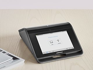 A Crestron tabletop device with a touchscreen interface, offering options for presenting sources via HDMI or wireless AirMedia connectivity.