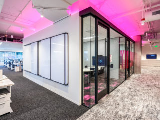 Modern office with glass-walled meeting rooms, pink accent lighting, a whiteboard wall, open workstations, and carpeted floors creating a vibrant workspace