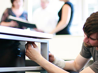 Technician repairing office furniture in a professional workspace with colleagues in the background, highlighting maintenance and support services.
