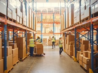 Warehouse scene with tall shelves stacked with boxes, workers in PPE organizing and inspecting packages, and forklifts in a large aisle.