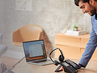 A professional using Poly technology, featuring a laptop, headset, and phone system, set on a desk in a modern workspace with soft furnishings.