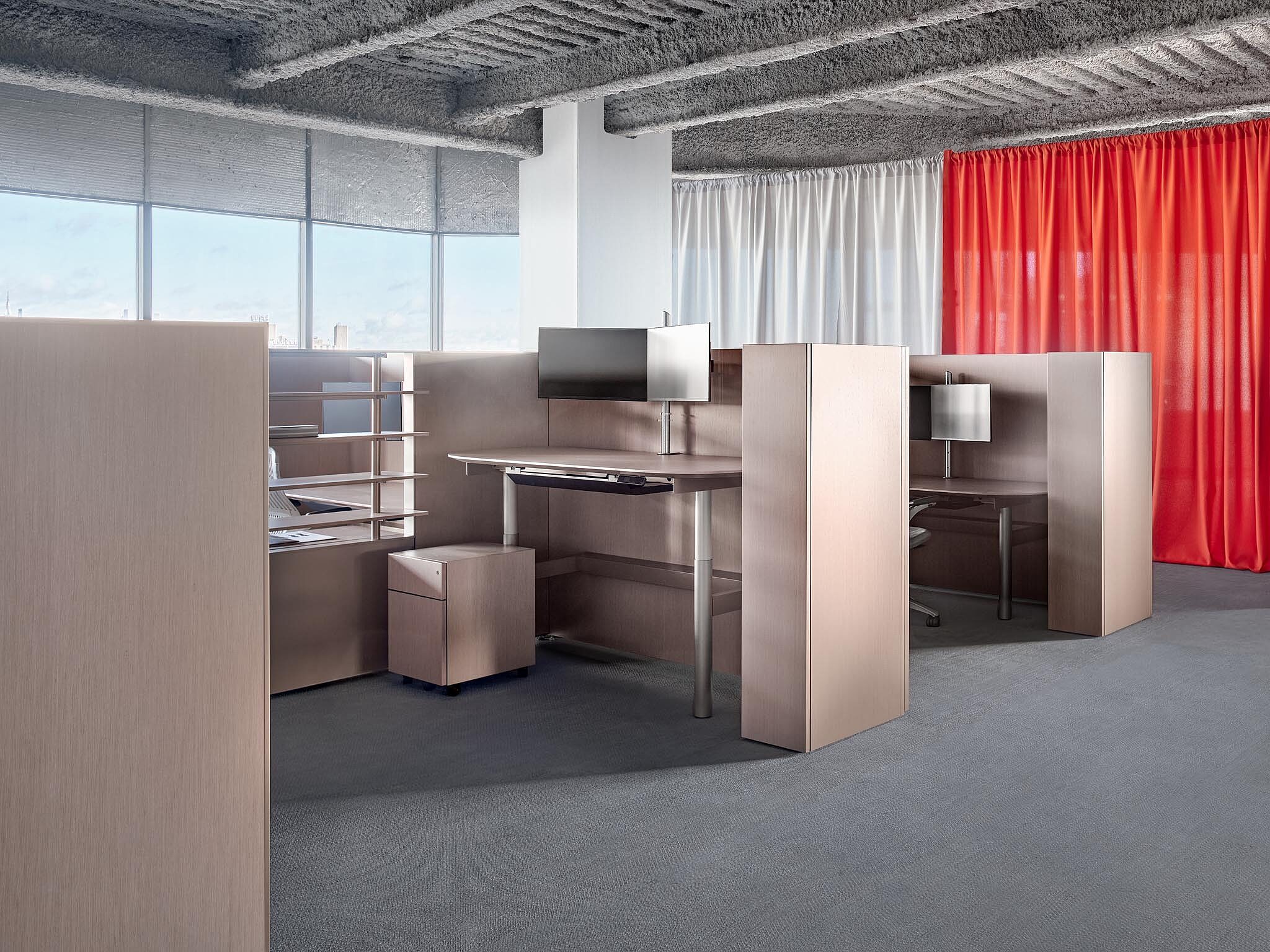 Modern office workstations with wood panels, monitors, and red curtain backdrop near large windows.