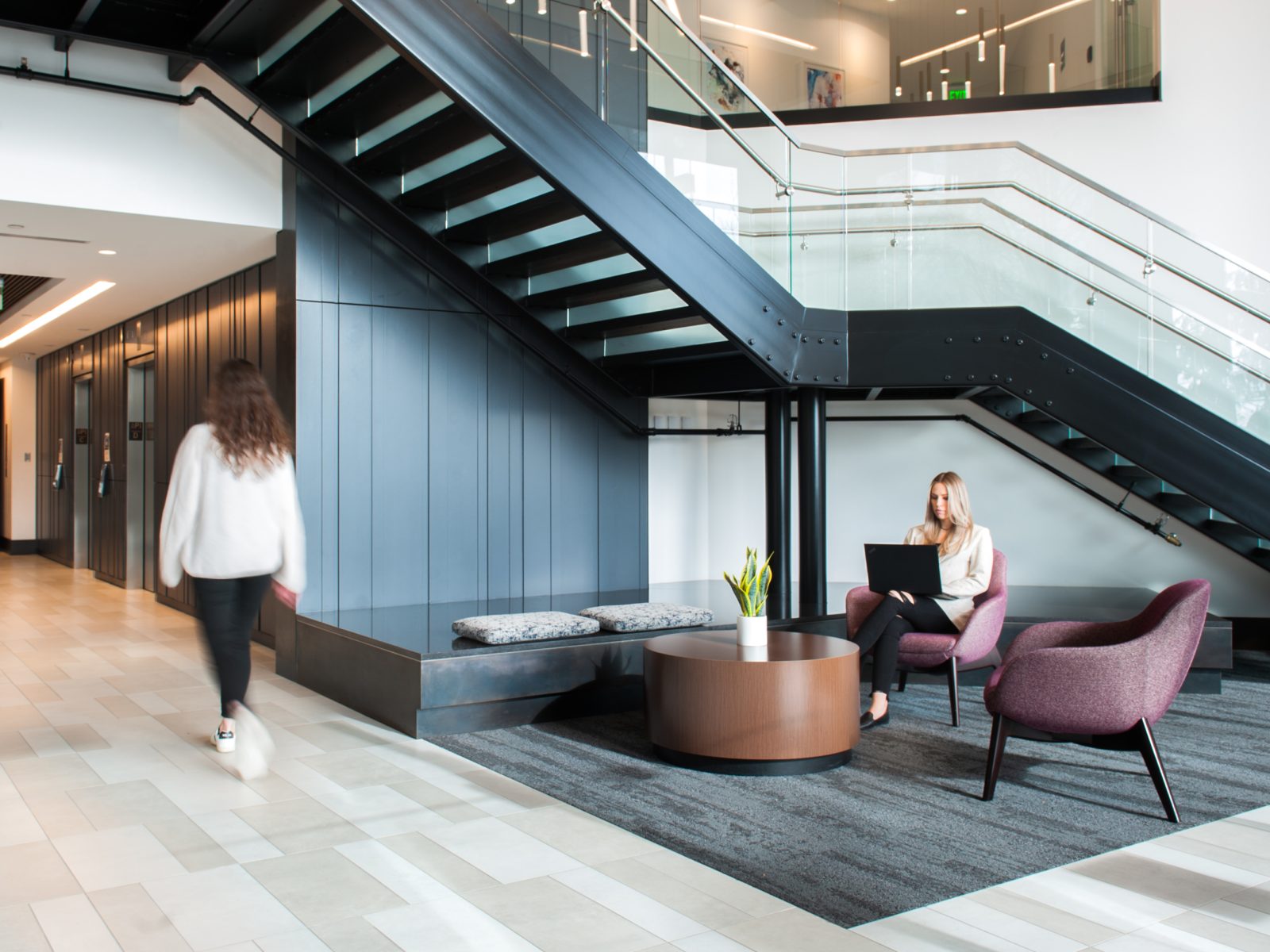 Modern office lobby with a black staircase, glass railings, and seating area. A woman works in a purple chair while another walks past on tiled floors.