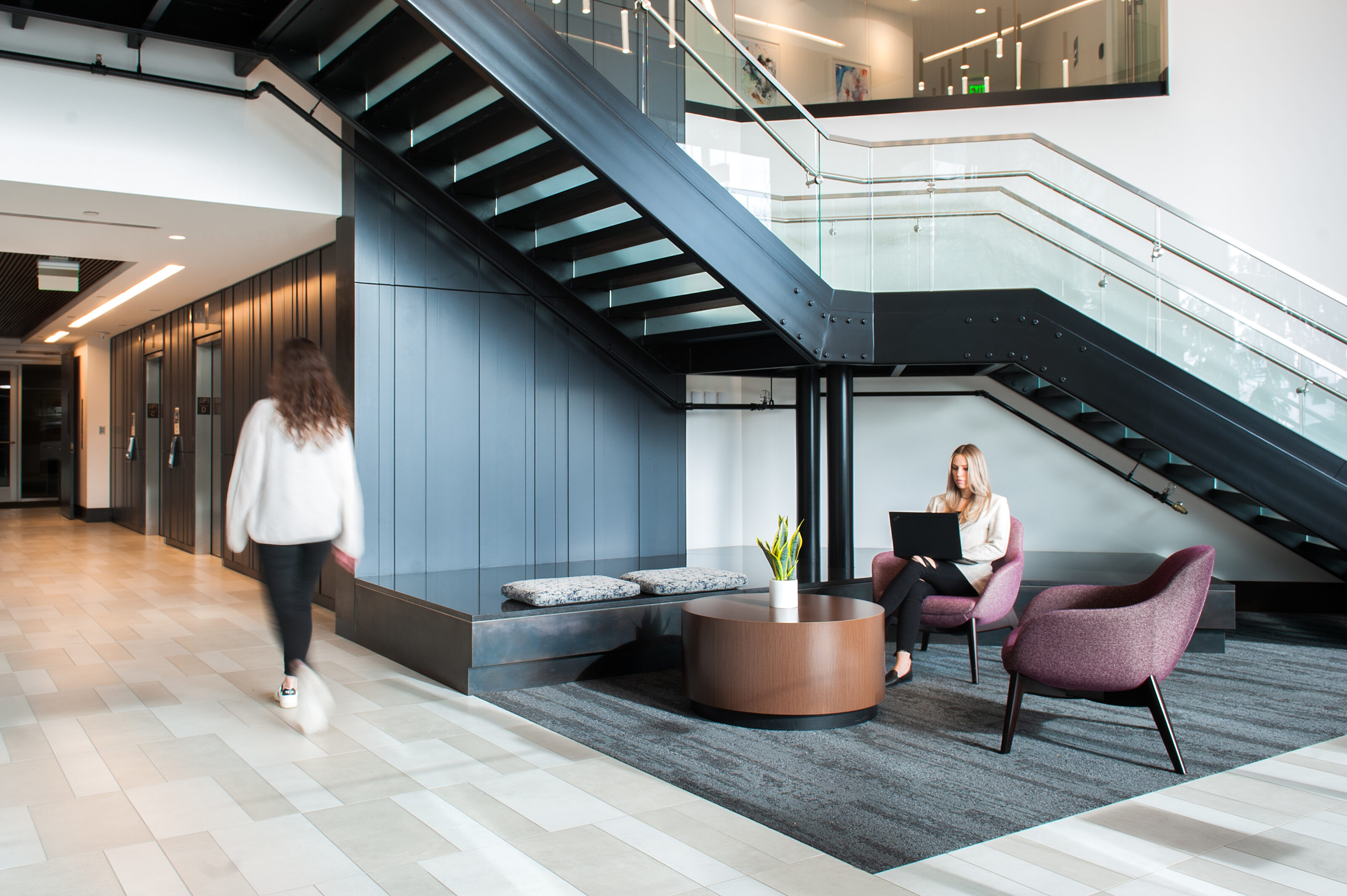 Modern office lobby with a black staircase, glass railings, and seating area. A woman works in a purple chair while another walks past on tiled floors.