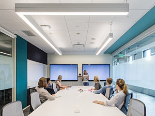 A group of people participates in a meeting in a modern conference room, featuring a long white table, glass walls, and dual screens for presentations.
