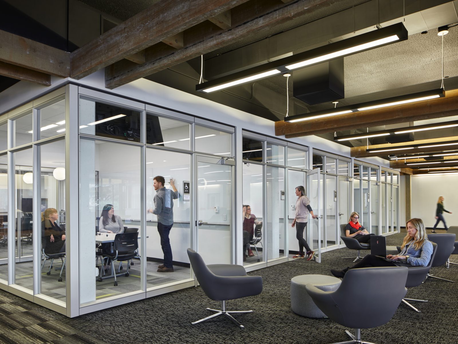 Modern office with glass-enclosed meeting rooms and open lounge seating. Employees collaborate in rooms, while others work on laptops in gray chairs.