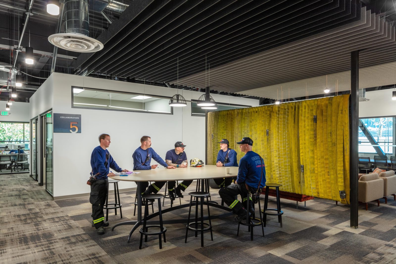 Modern workspace with a high-top table, stools, and five firefighters in blue uniforms, with yellow fire hoses serving as a room divider.