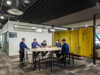 Modern workspace with a high-top table, stools, and five firefighters in blue uniforms, with yellow fire hoses serving as a room divider.