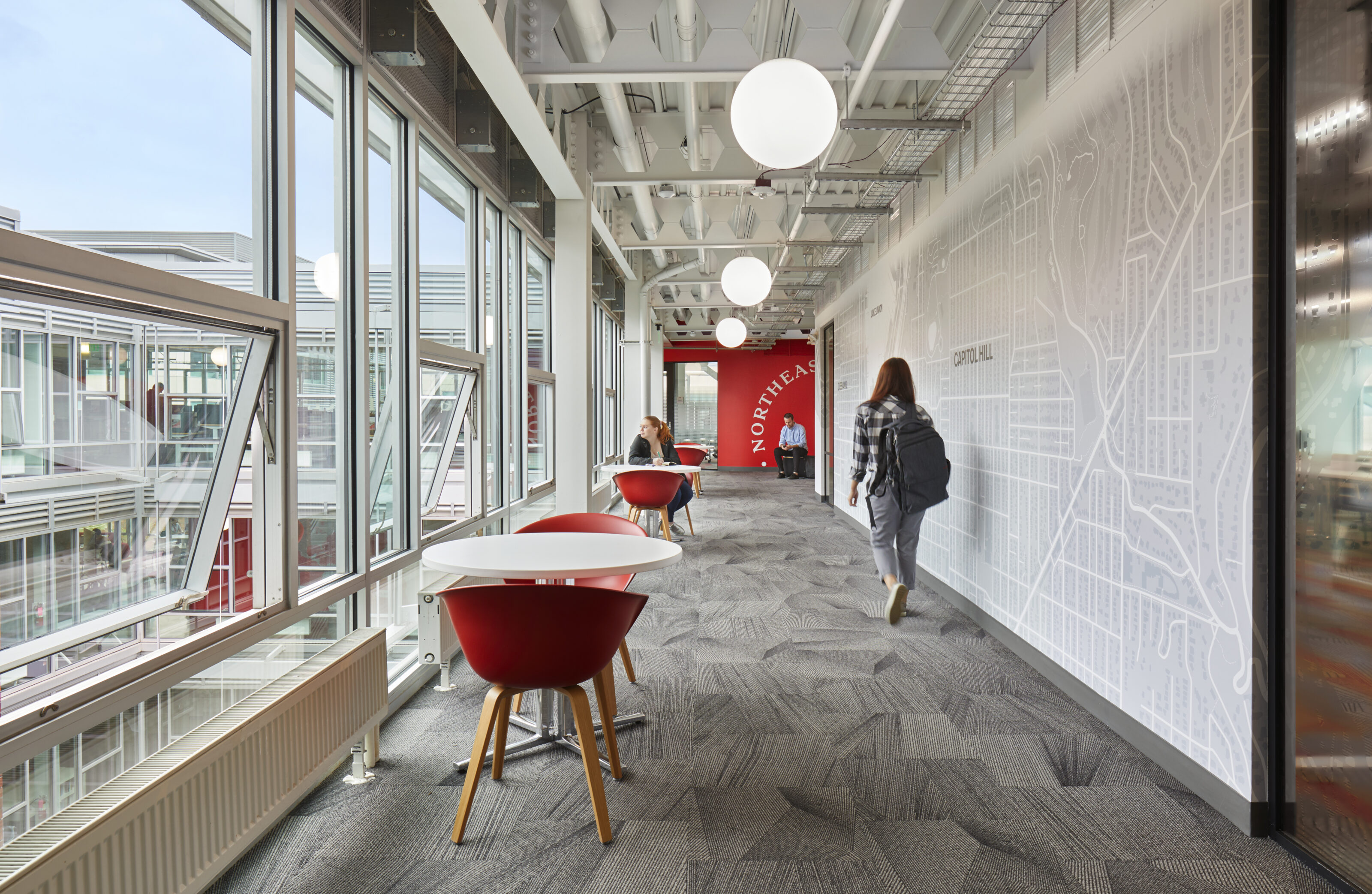 Bright office hallway with large windows, red about a chairs at white everywhere tables, gray carpeting, map-themed wall art, and people walking.