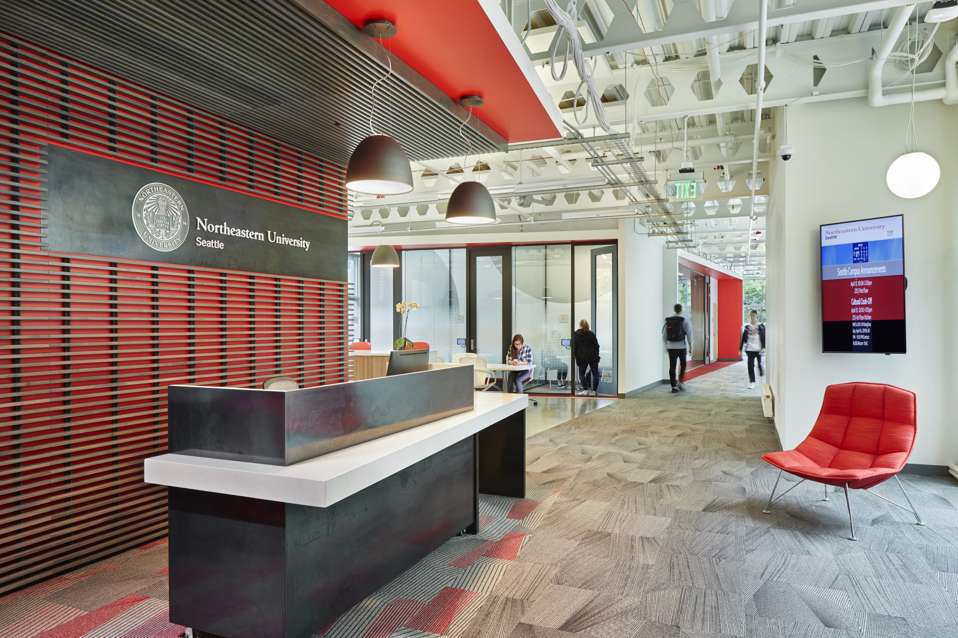 Northeastern University reception area with a modern desk, red accent wall, pendant lighting, seating, and a hallway leading to glass-walled meeting rooms