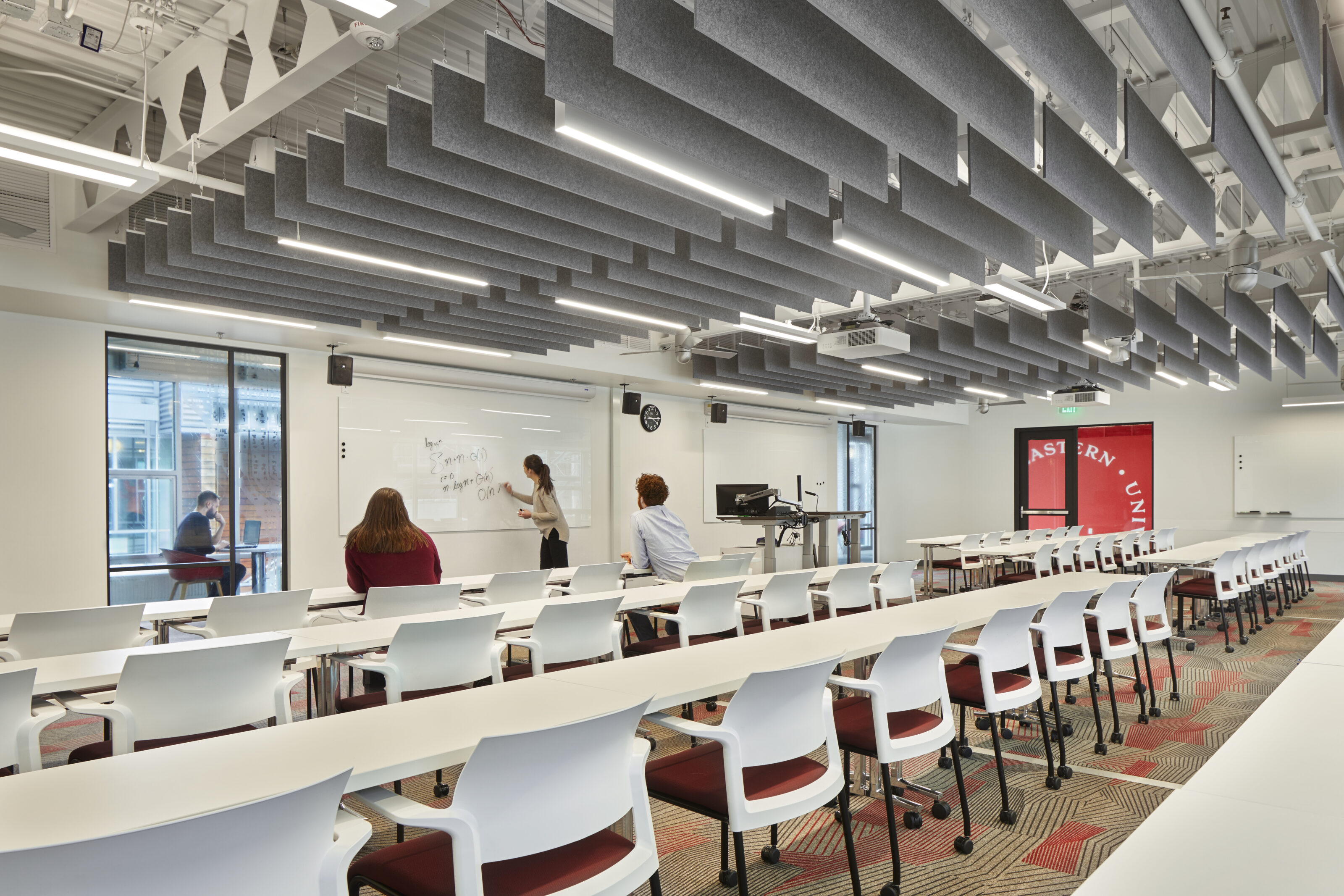 University classroom with rows of white desks, red-accented chairs, acoustic ceiling panels, and a professor writing on a whiteboard while students observe