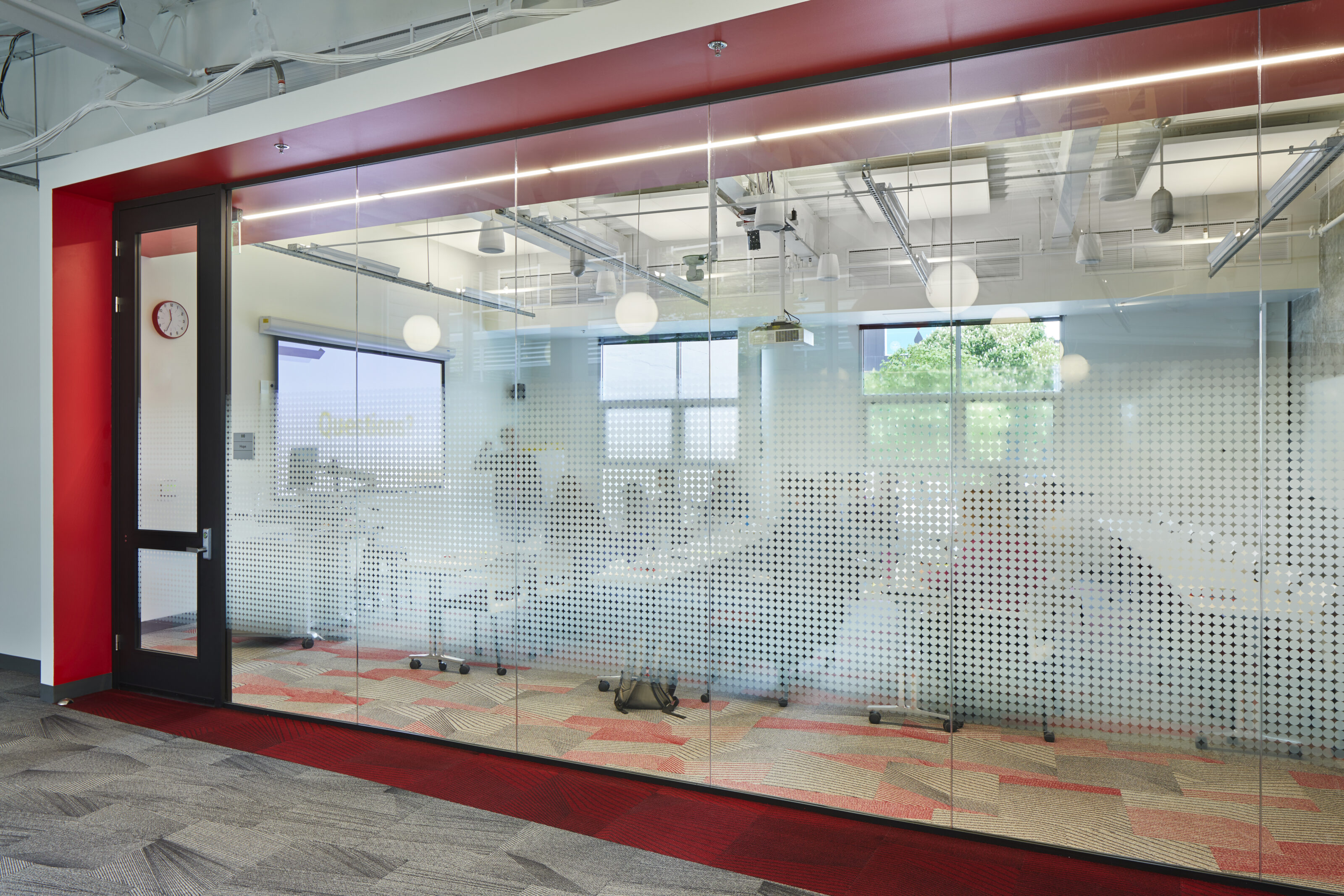 Glass-walled classroom with frosted designs, red accents, and modern lighting. Inside, a projector screen and desks are visible in the bright, open space.