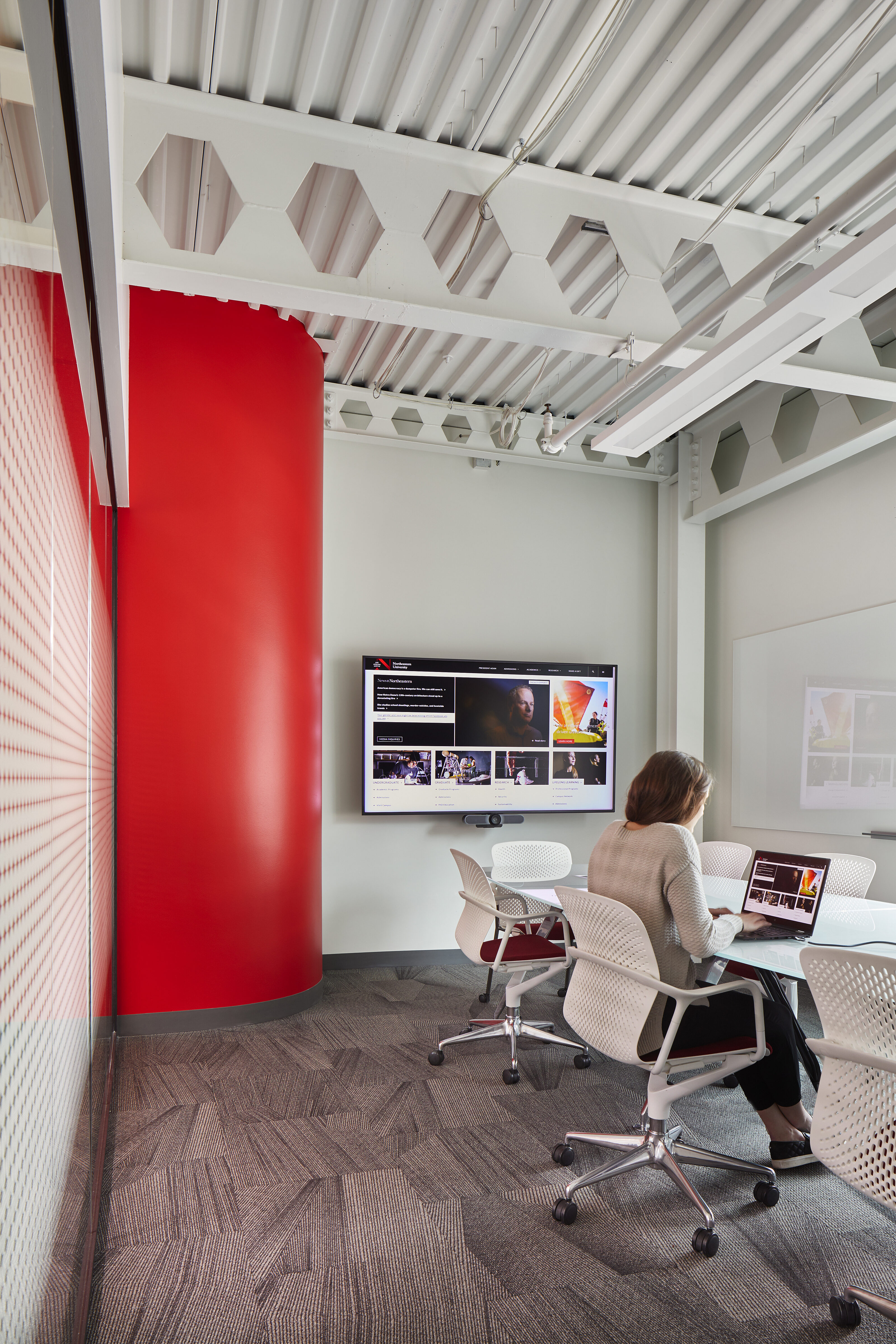 Modern meeting space with white chairs, a red accent wall, and a wall-mounted screen. A person works on a laptop at a glass table under exposed ceilings.