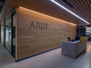 Modern reception area featuring a wood-paneled wall with "ARUP" signage, a sleek concrete desk, and a ceiling with wood slats and integrated lighting.