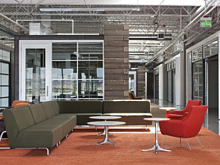 Modern office lounge with brown sectional seating, red chairs, and round tables on orange carpet. Glass walls and exposed ceilings create an open feel.