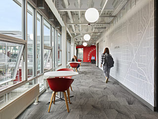 Bright office hallway with large windows, red about a chairs at white everywhere tables, gray carpeting, map-themed wall art, and people walking.