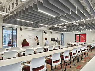 University classroom with rows of white desks, red-accented chairs, acoustic ceiling panels, and a professor writing on a whiteboard while students observe